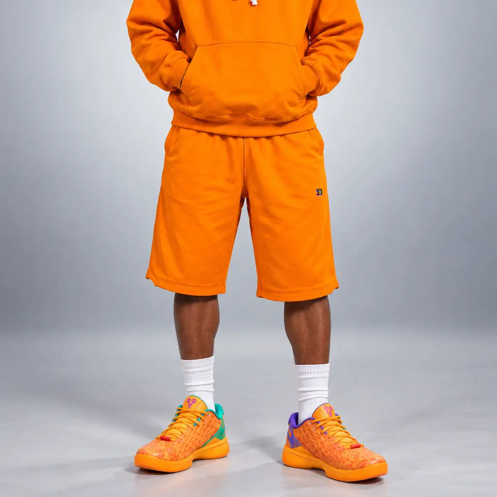 A model stands against a gray studio background wearing a matching bright orange hoodie and shorts set, white crew socks, and orange sneakers with small multicolor accents.