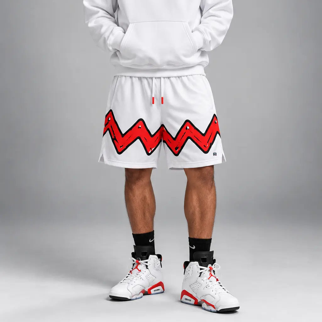 A male model wearing a white pullover hoodie and matching white shorts with a bold red and black zigzag graphic across the shorts, styled with black socks and white sneakers, against a gray studio background.