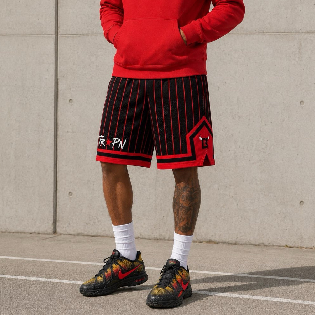 Person wearing red hoodie, black and red striped shorts, and colorful sneakers against a concrete wall.