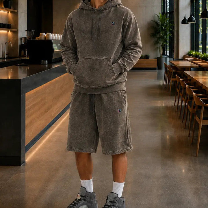 A man wearing a matching gray corduroy hoodie and shorts set stands in a modern, spacious cafe interior with wood tables, large windows, and concrete floors.