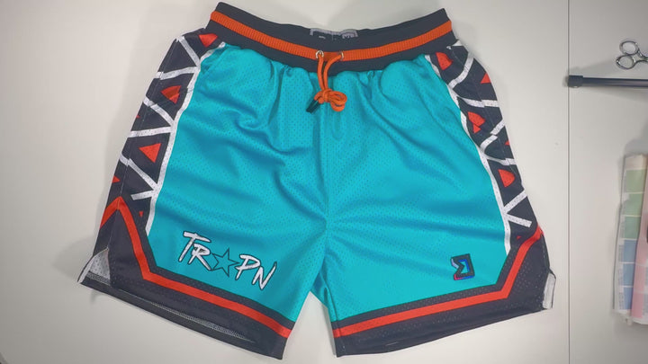 Trap Star Mesh Pro basketball shorts in aqua blue with tribal-style black/orange detailing, 2-inch pro waistband, and signature TR✶PN script by Trap’N Shorts.